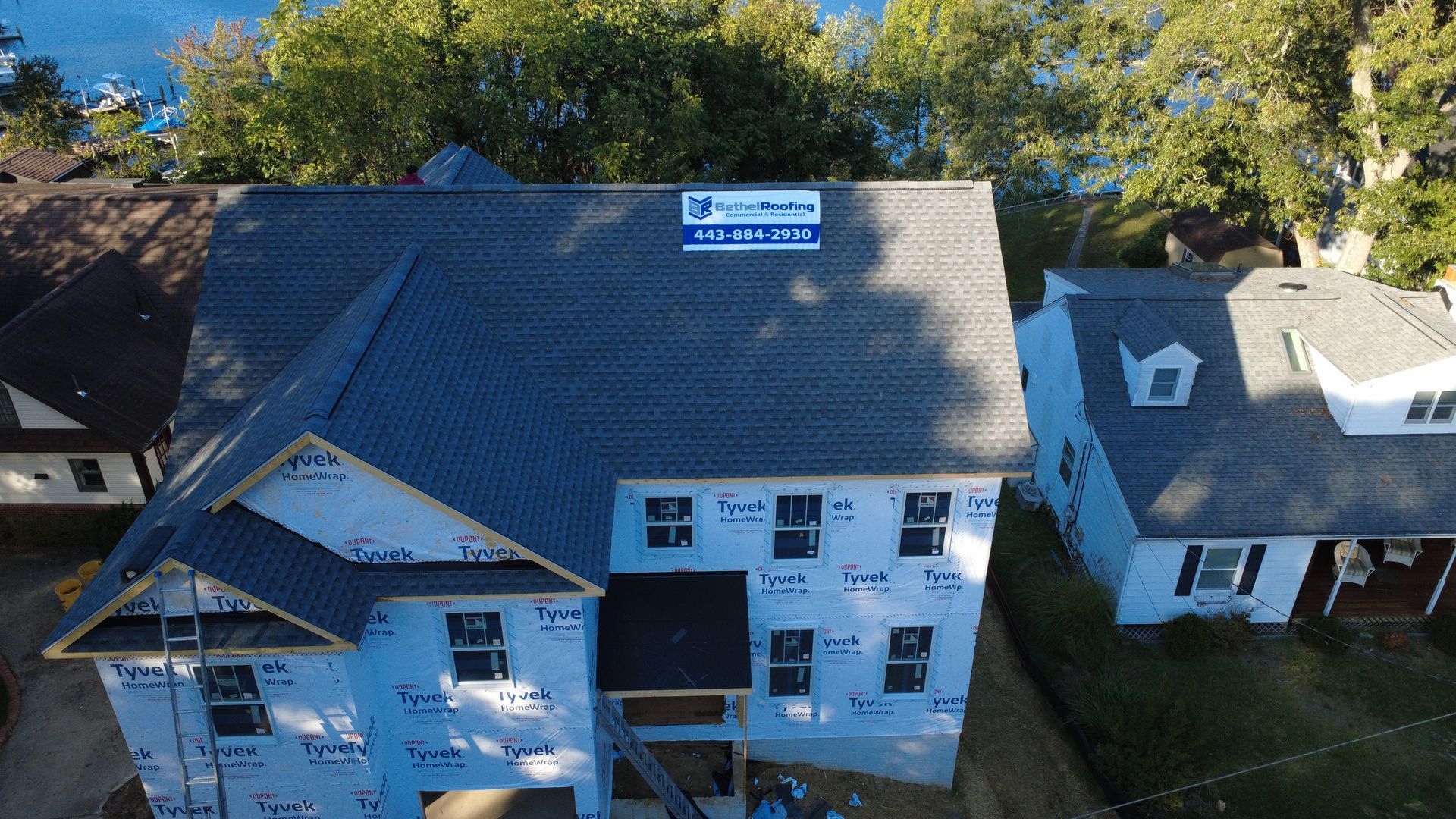 New house under construction with dark gray roof, blue wrap, and partially framed, with a lake in the background.