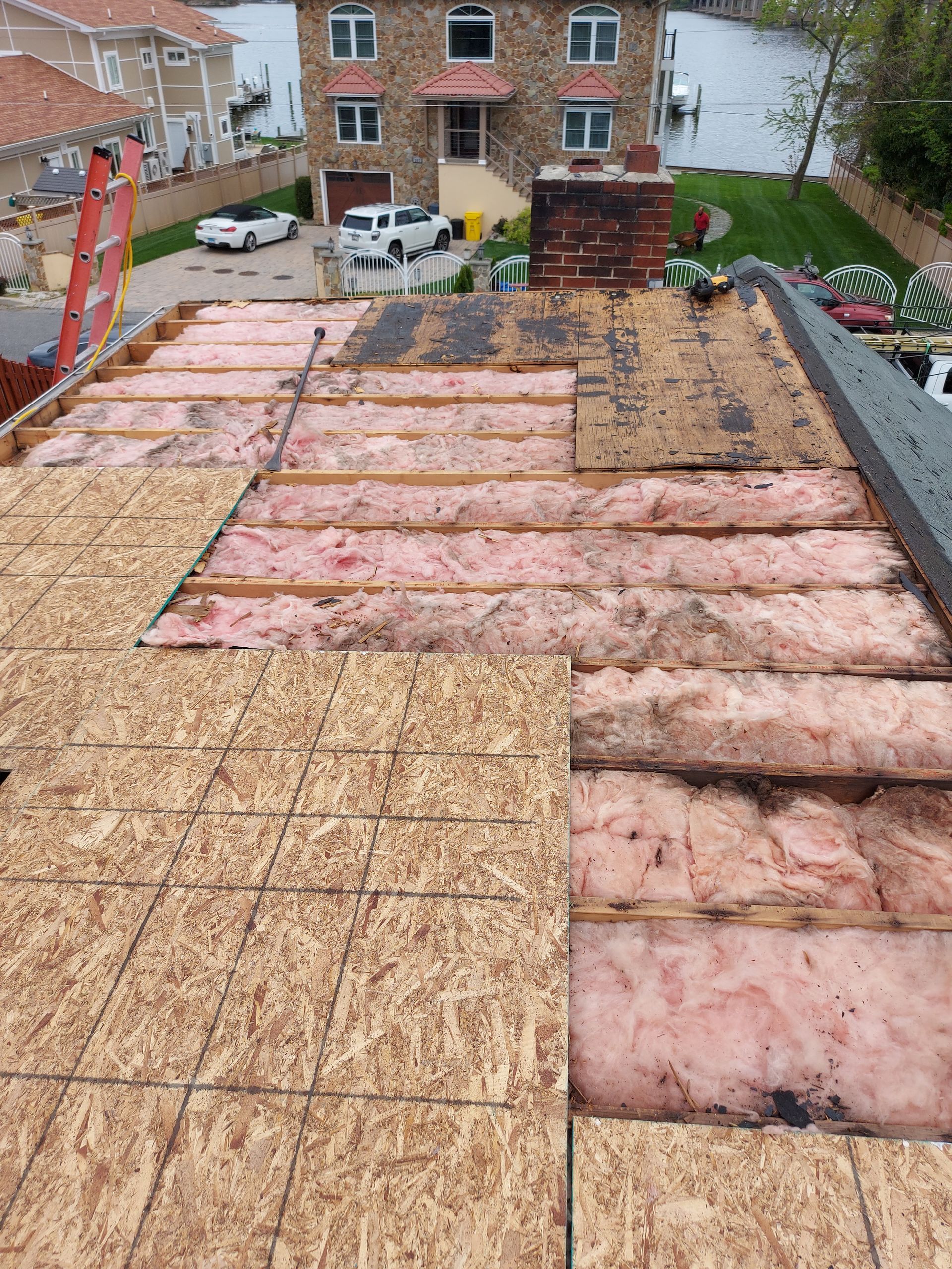 Roof partially stripped during repair, showing insulation and plywood. Houses and water in the background.