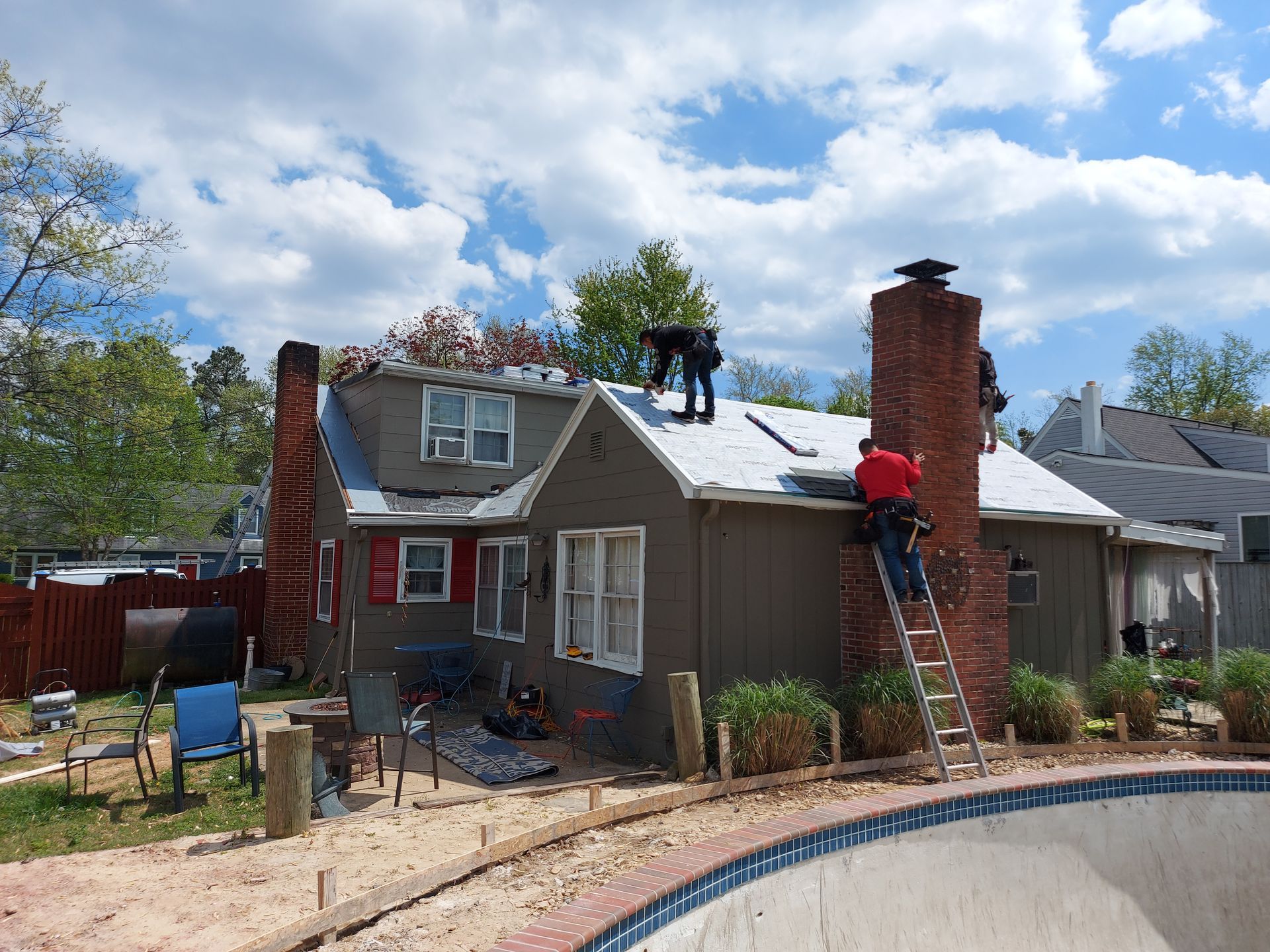 Workers replacing a roof on a small house with a brick chimney. People on roof and ladder. Pool in foreground.