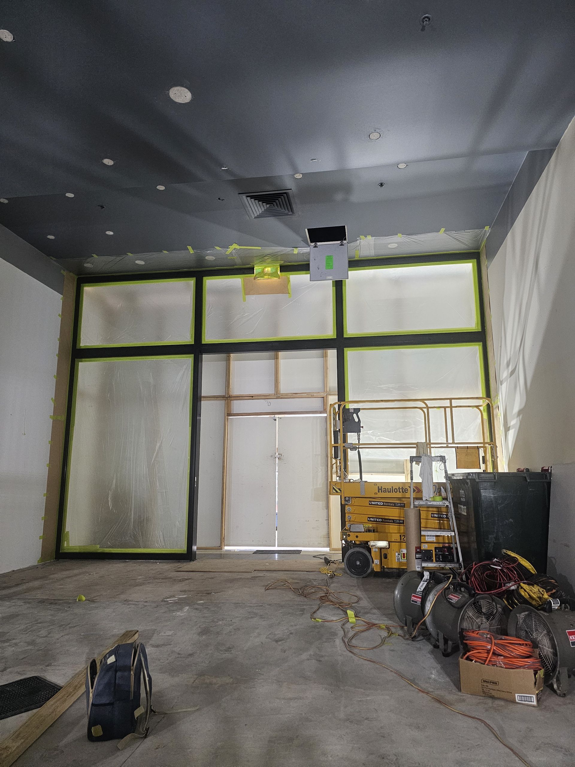 Construction in progress: Storefront with taped glass, open doorway, yellow lift, gray ceiling, concrete floor — Prestige Painting NT in Zuccoli, NT
