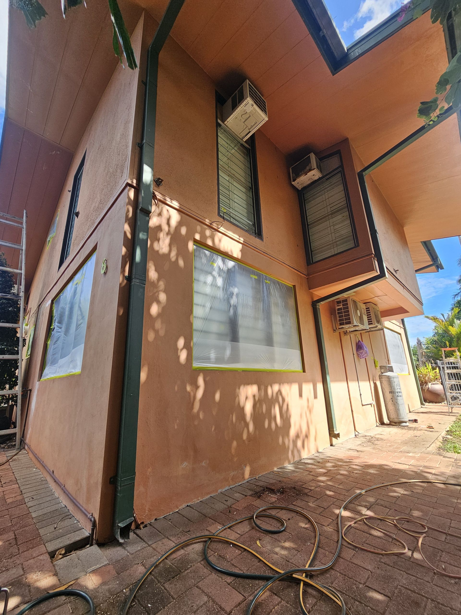Tan building exterior with green trim and AC units. Windows are taped, brick patio, sunny — Prestige Painting NT in Zuccoli, NT