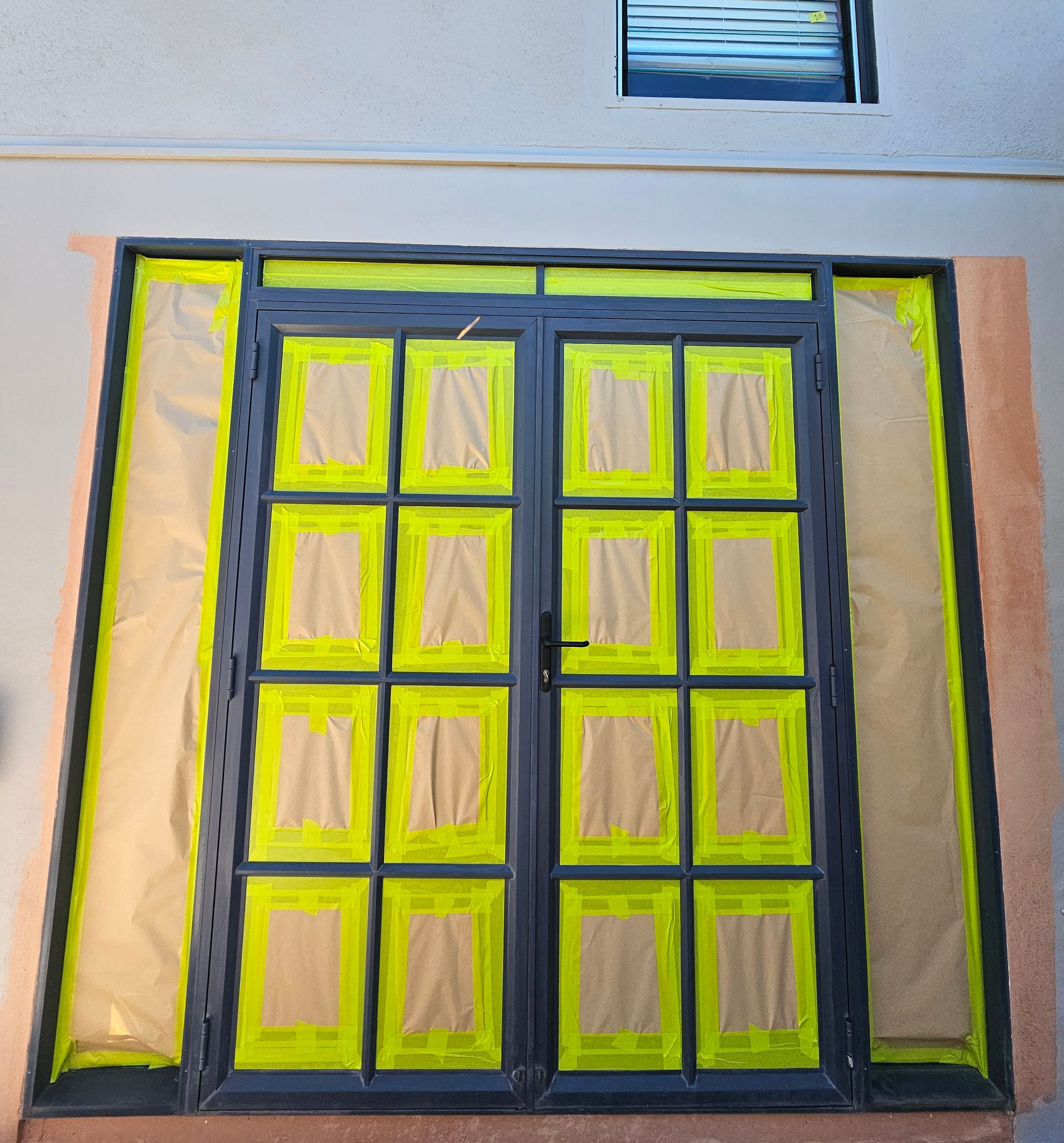 Black-framed double doors with multiple panes, yellow tape, and protective covering. In a building, with a window visible above — Prestige Painting NT in Zuccoli, NT