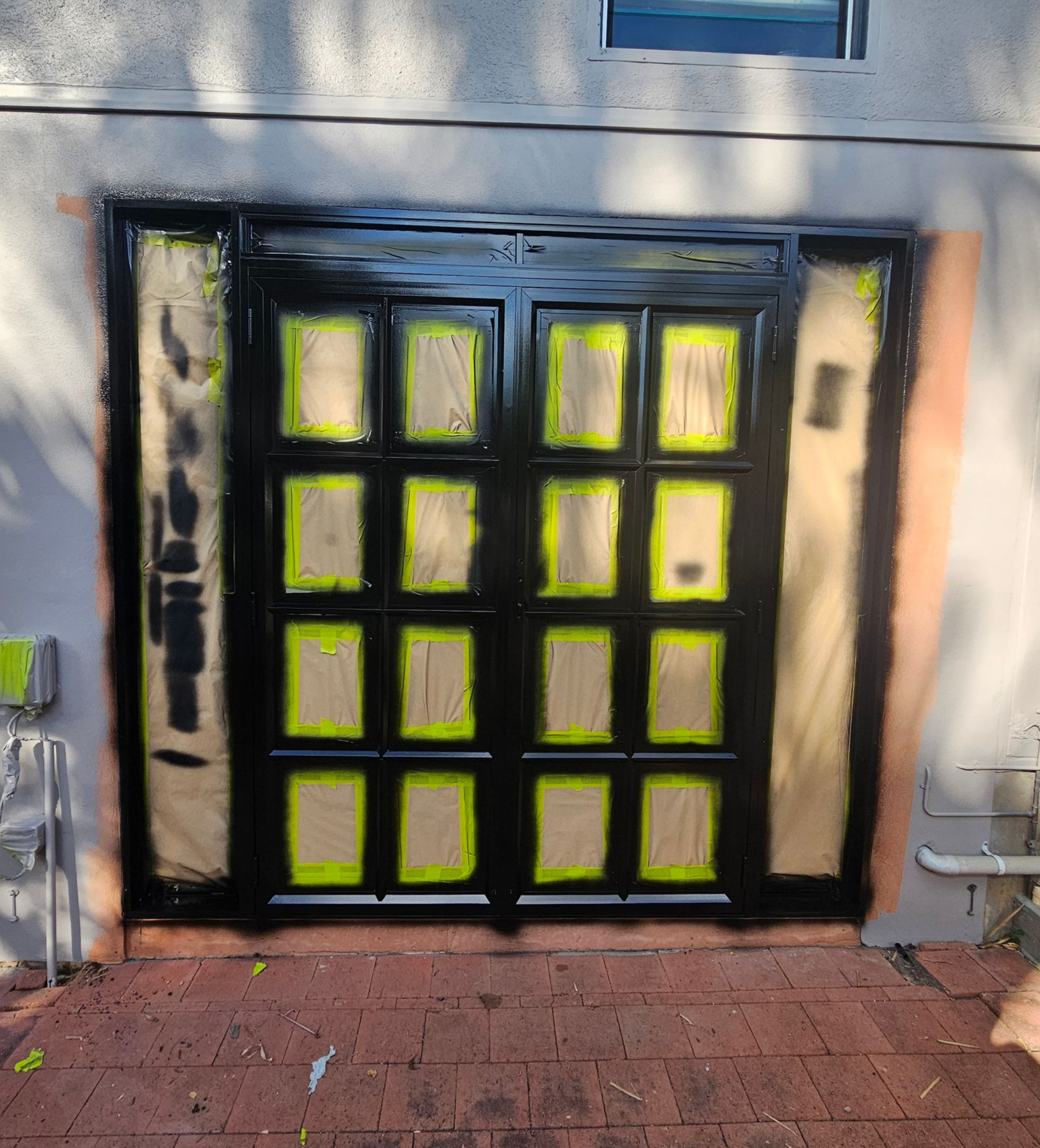 Black-painted double doors with window panes, taped with yellow, are being painted on a light-coloured building with red brick — Prestige Painting NT in Zuccoli, NT