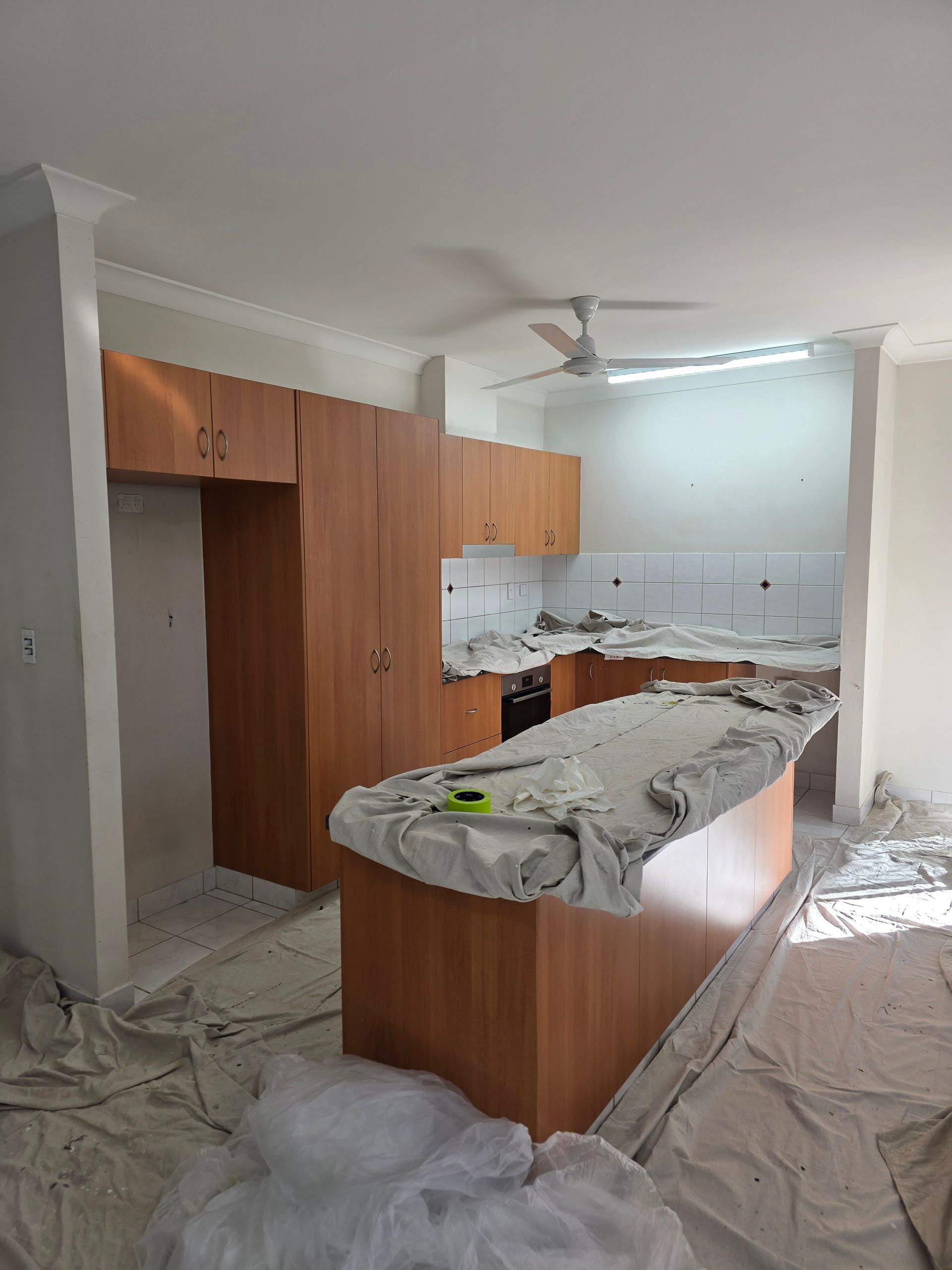 Kitchen undergoing renovation; light brown cabinets, island, countertops covered, white walls, and ceiling fan — Prestige Painting NT in Zuccoli, NT