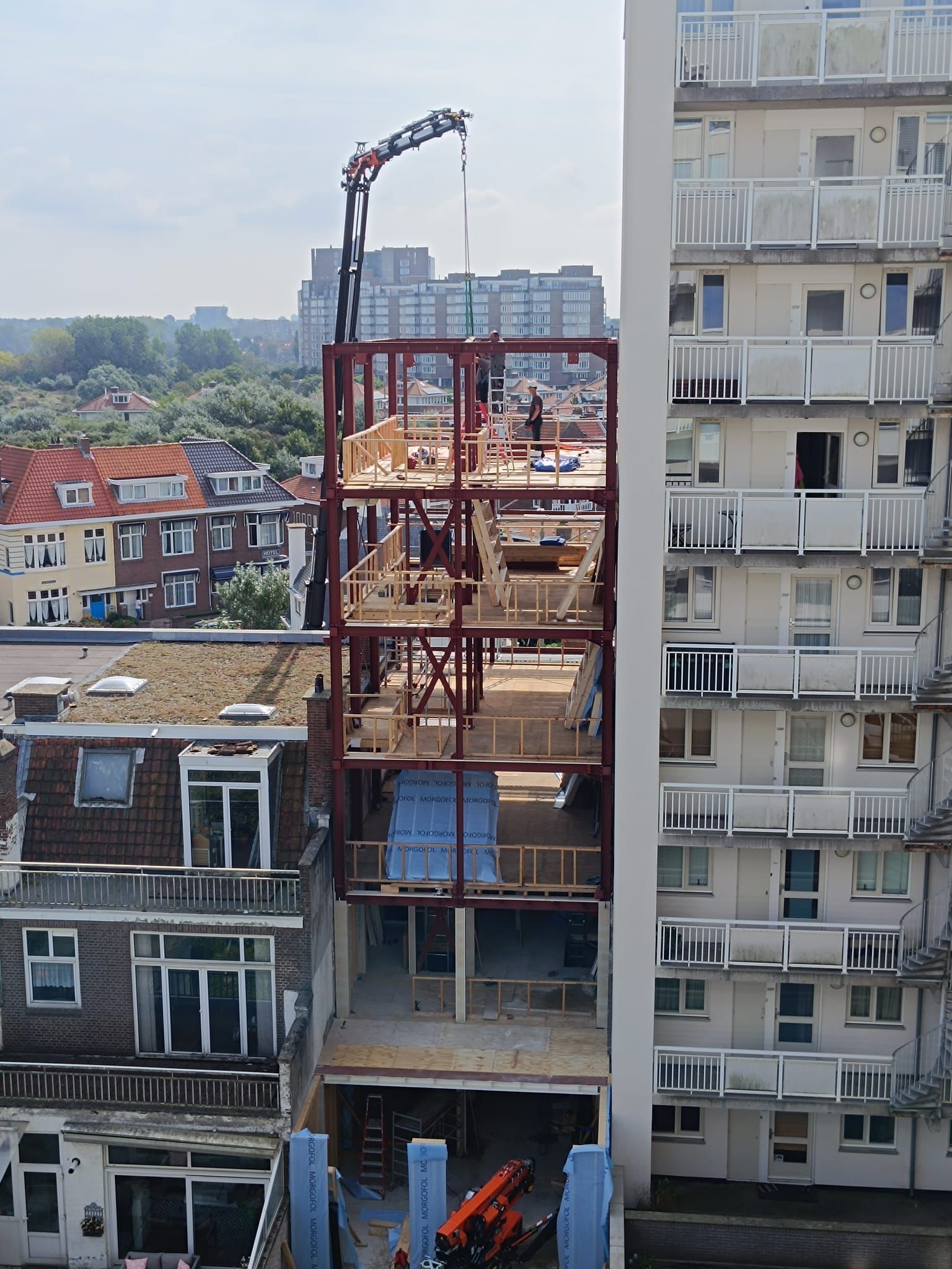 De constructie van een geraamte voor een meerlaags gebouw tussen bestaande structuren; een kraan hijst materialen omhoog.