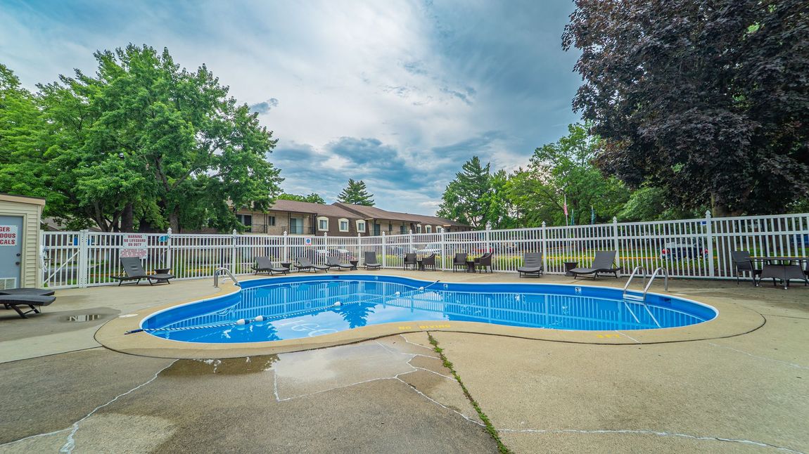 Swimming pool surrounded by a white fence and concrete deck. Townhouses in background, cloudy sky overhead.