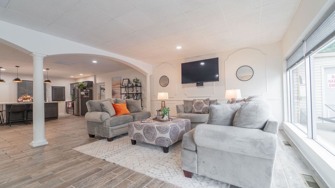 Living room with gray sofas, patterned ottoman, TV, and large windows. Archway leads to kitchen.