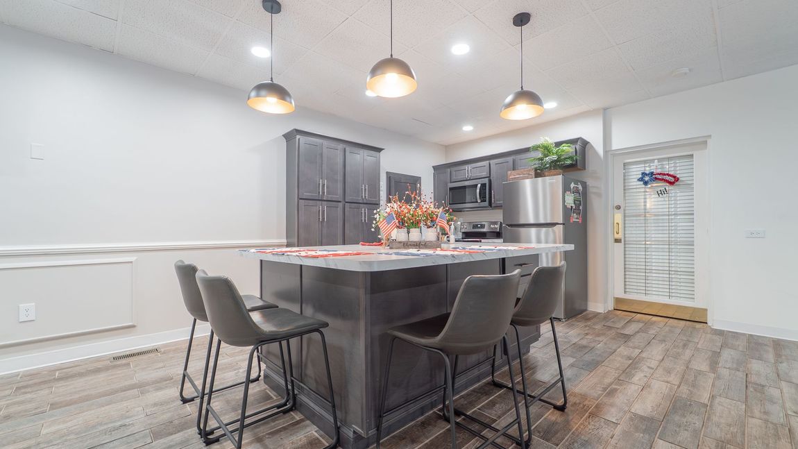 Kitchen with island, gray cabinets, stainless steel appliances, pendant lights, bar stools, and hardwood floors.