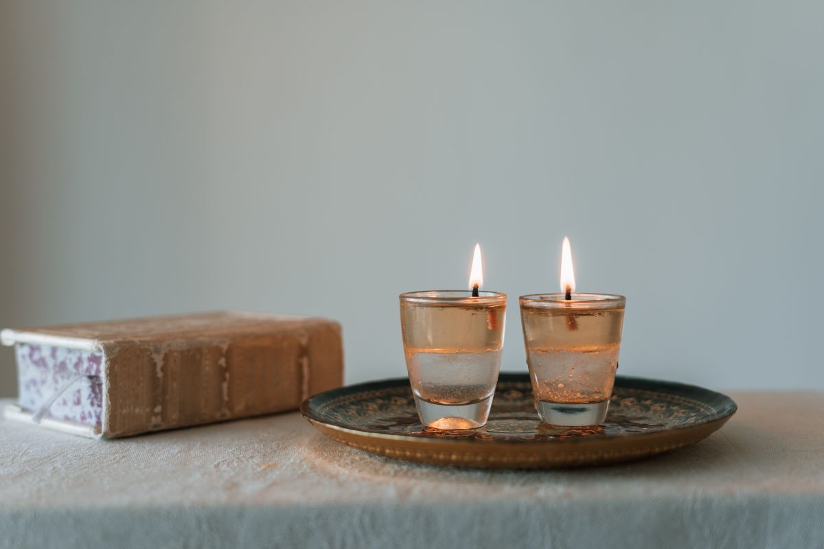 Two lit votive candles in glass holders sit on a patterned tray next to a vintage book on a light-colored surface.