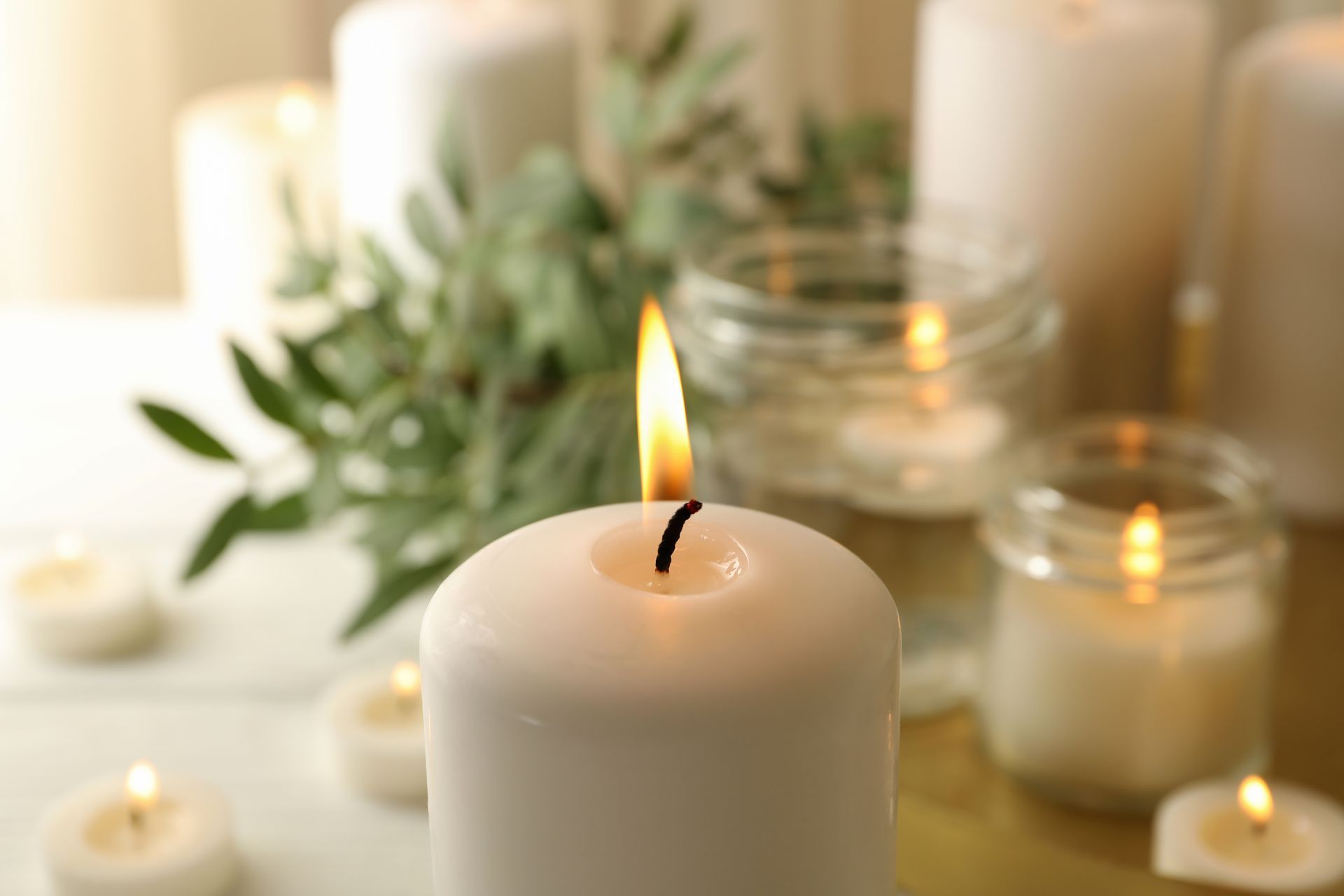 Lit white candle with soft bokeh of other candles and greenery in a warm, serene setting