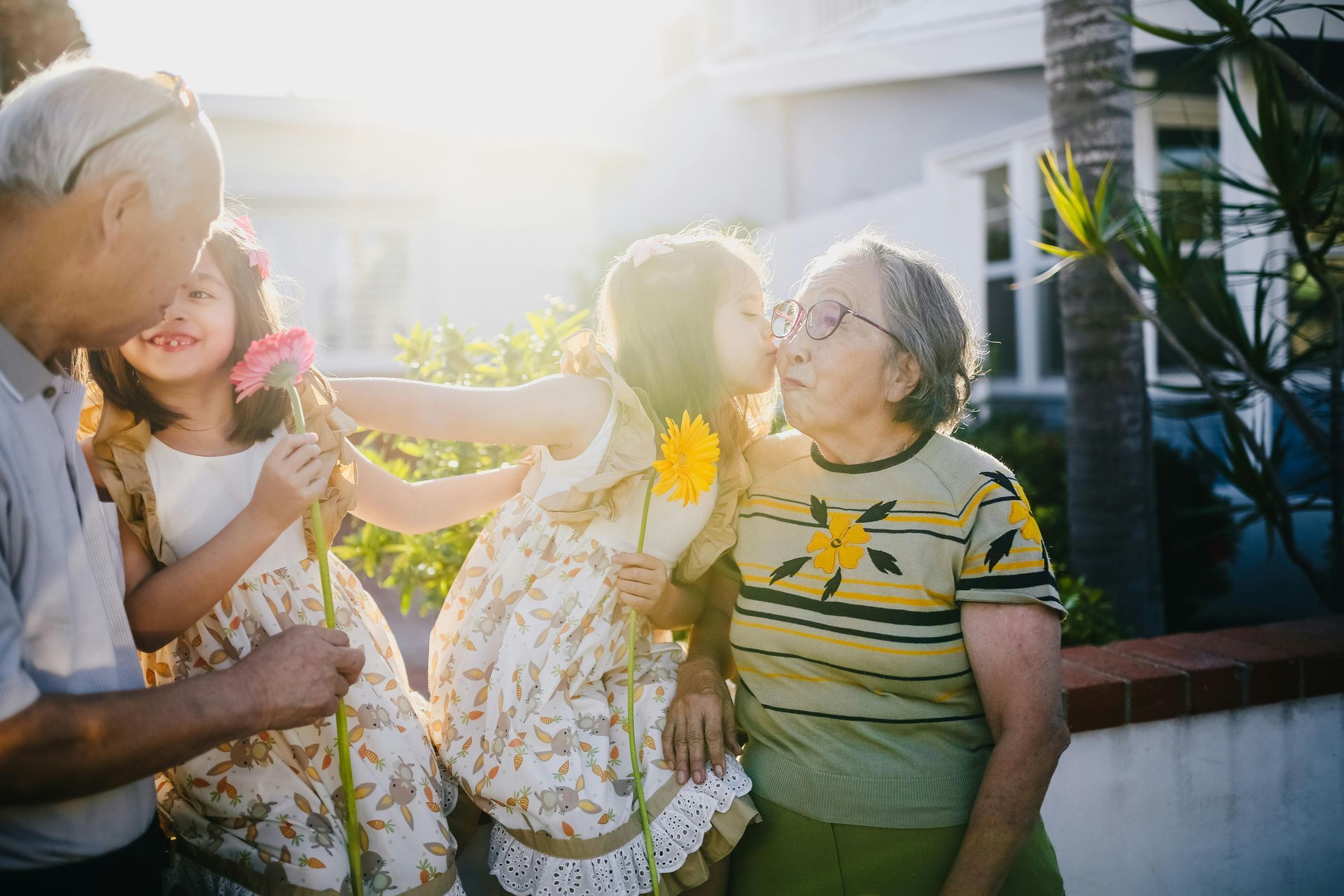 Two children and an adult laughing together in bright sunlight outdoors, holding flowers by a house.