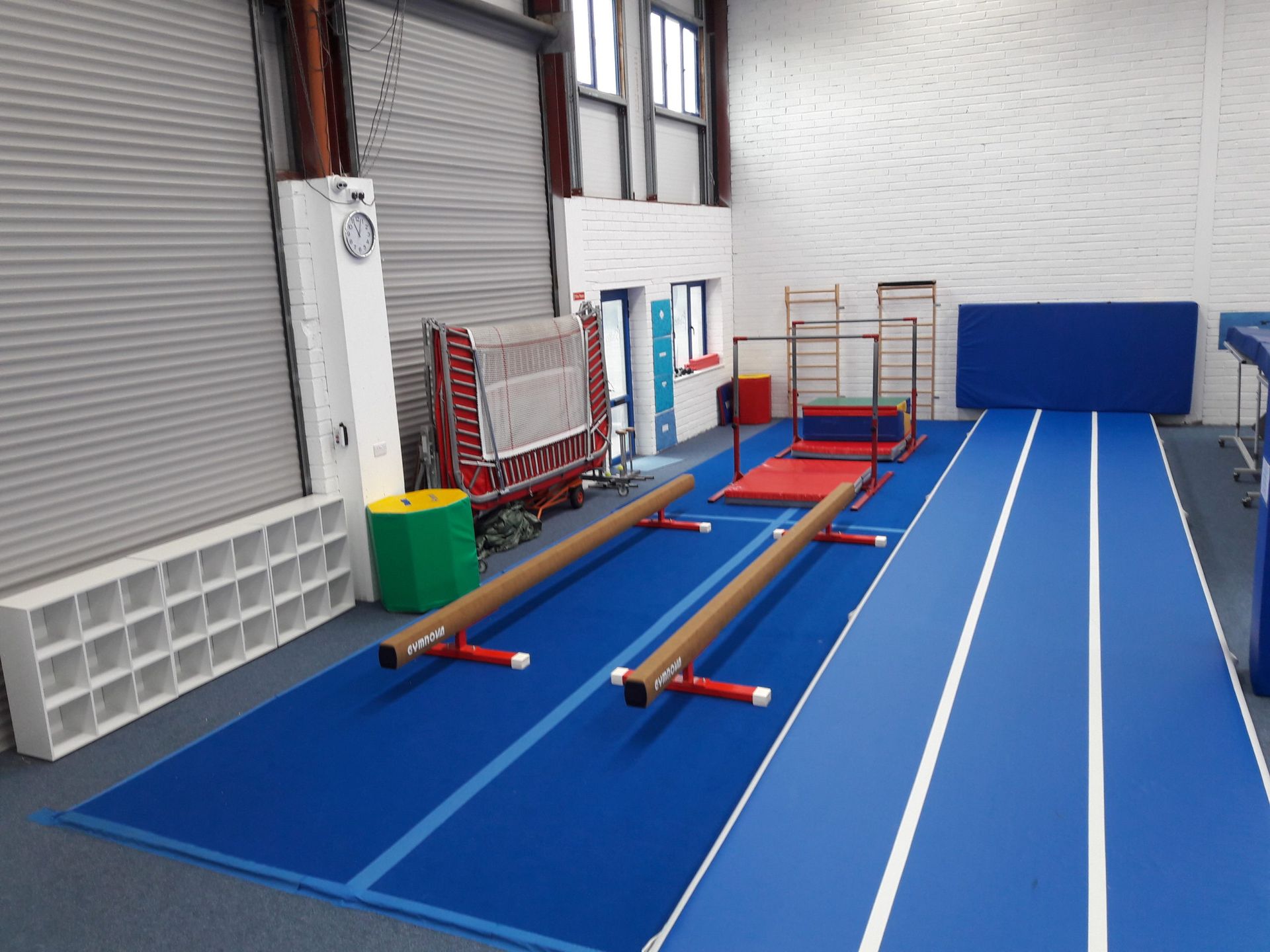Indoor gym with blue floor mats, balance beams, and training equipment against white walls and roll-up doors