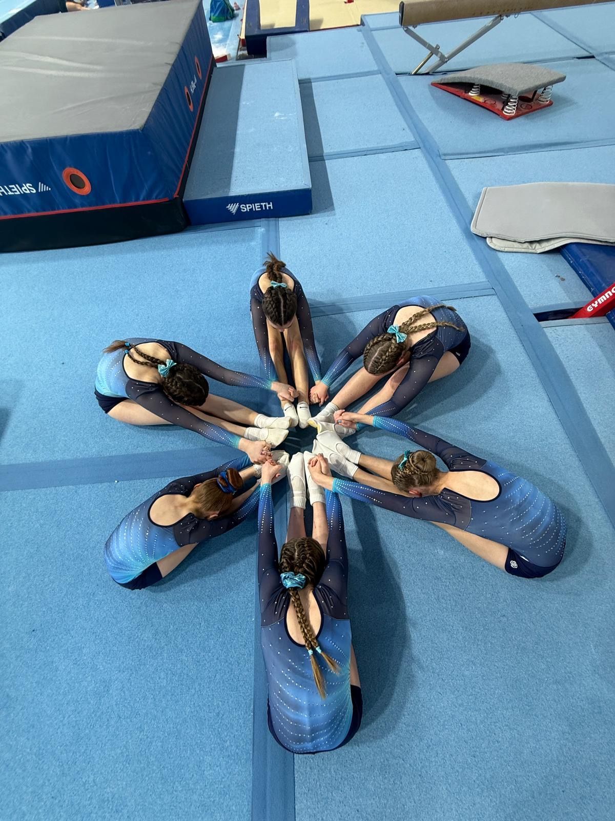 Gymnast in blue leotard performing a split leap on a balance beam under a stadium roof