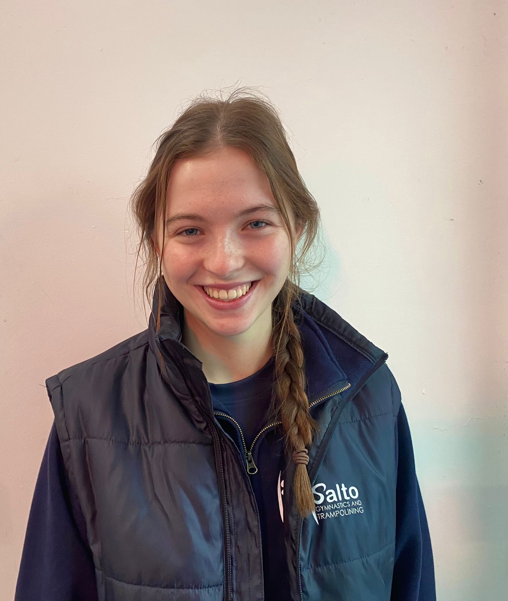 Smiling person in a navy vest with braided hair, standing against a plain light wall.