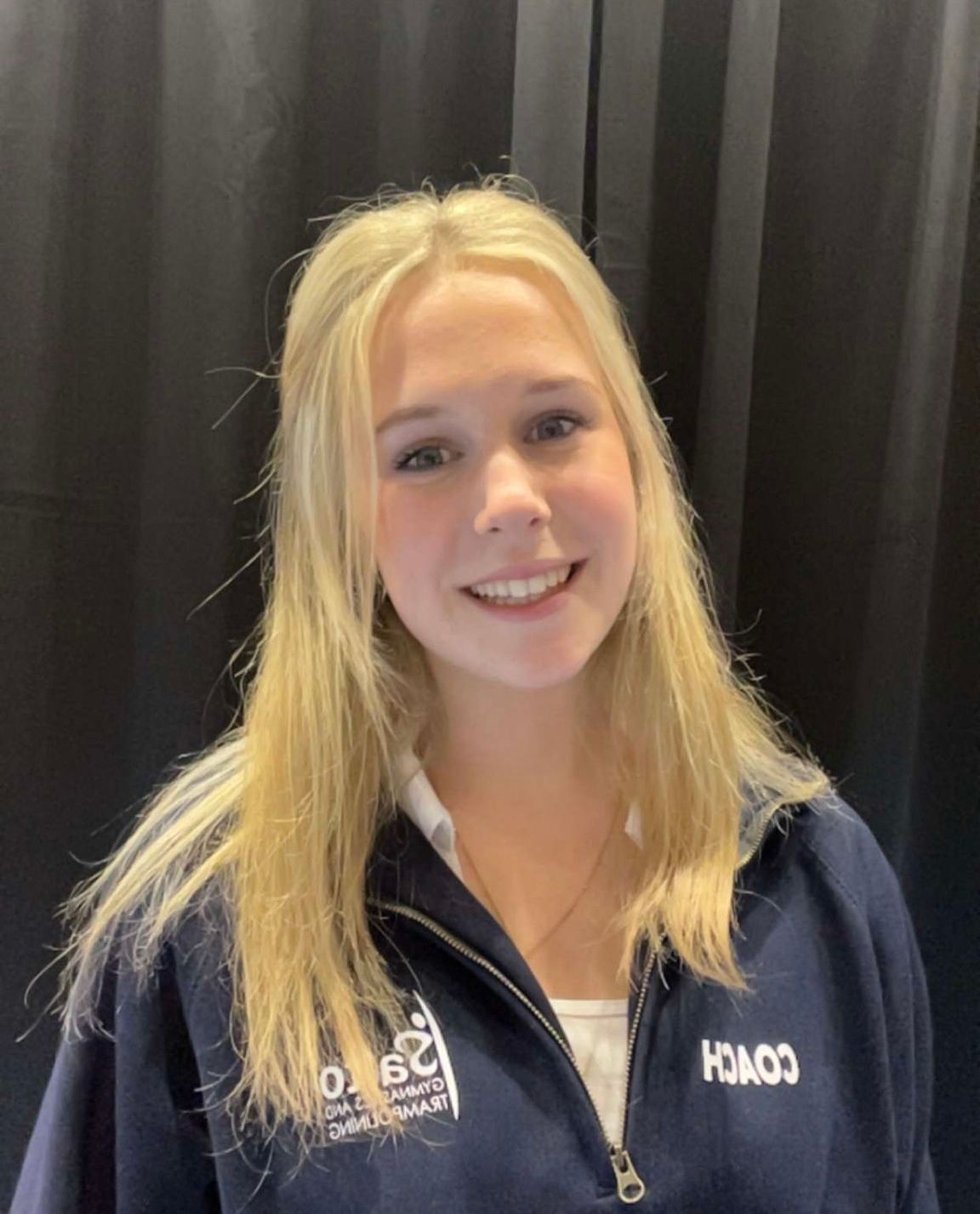 Smiling blonde woman in a navy jacket standing before black curtains.