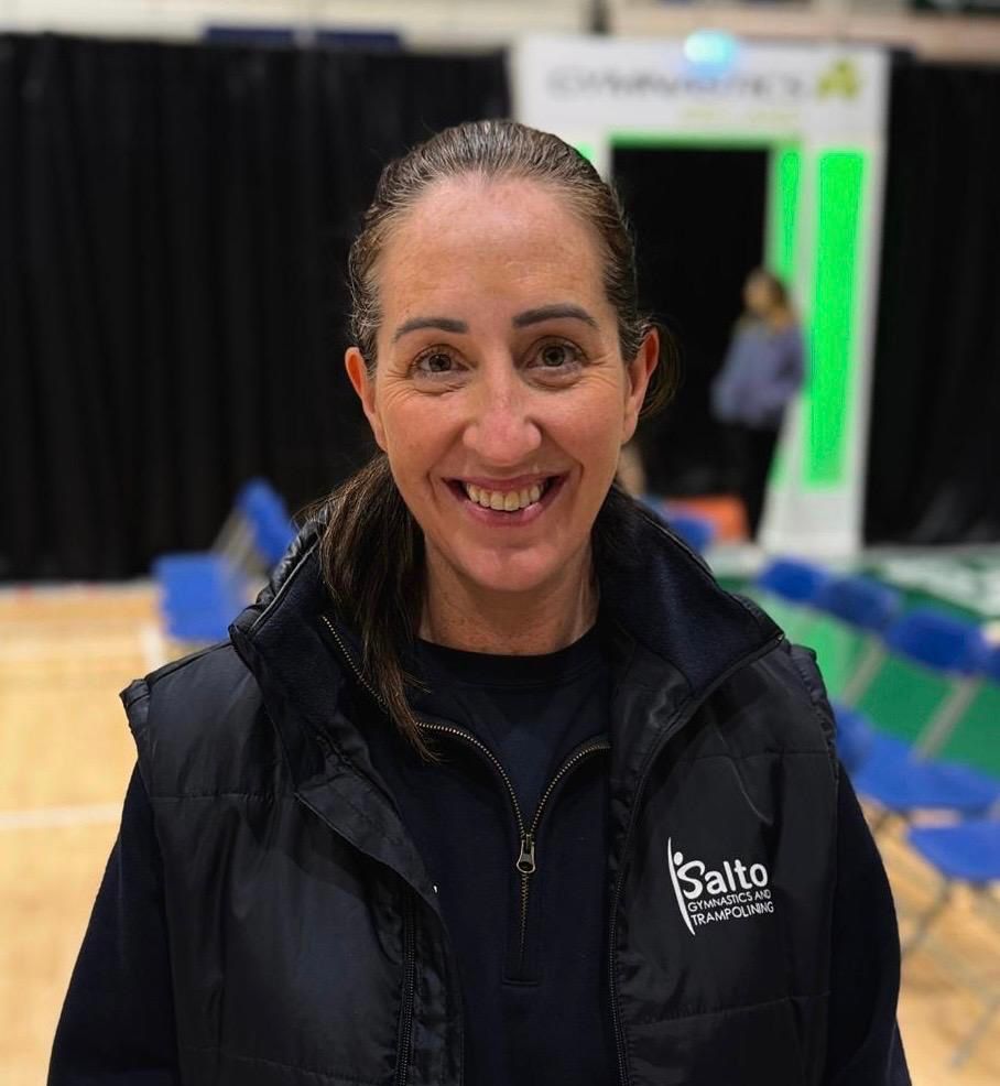 Smiling woman in a black jacket standing indoors at an event booth with blue chairs and a green-lit display.