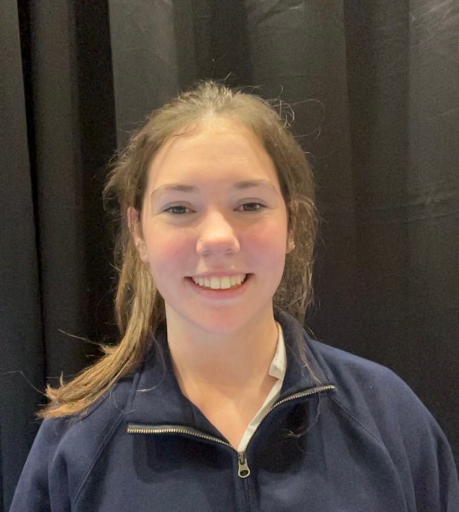 Smiling person in a navy zip-up jacket standing in front of black curtains