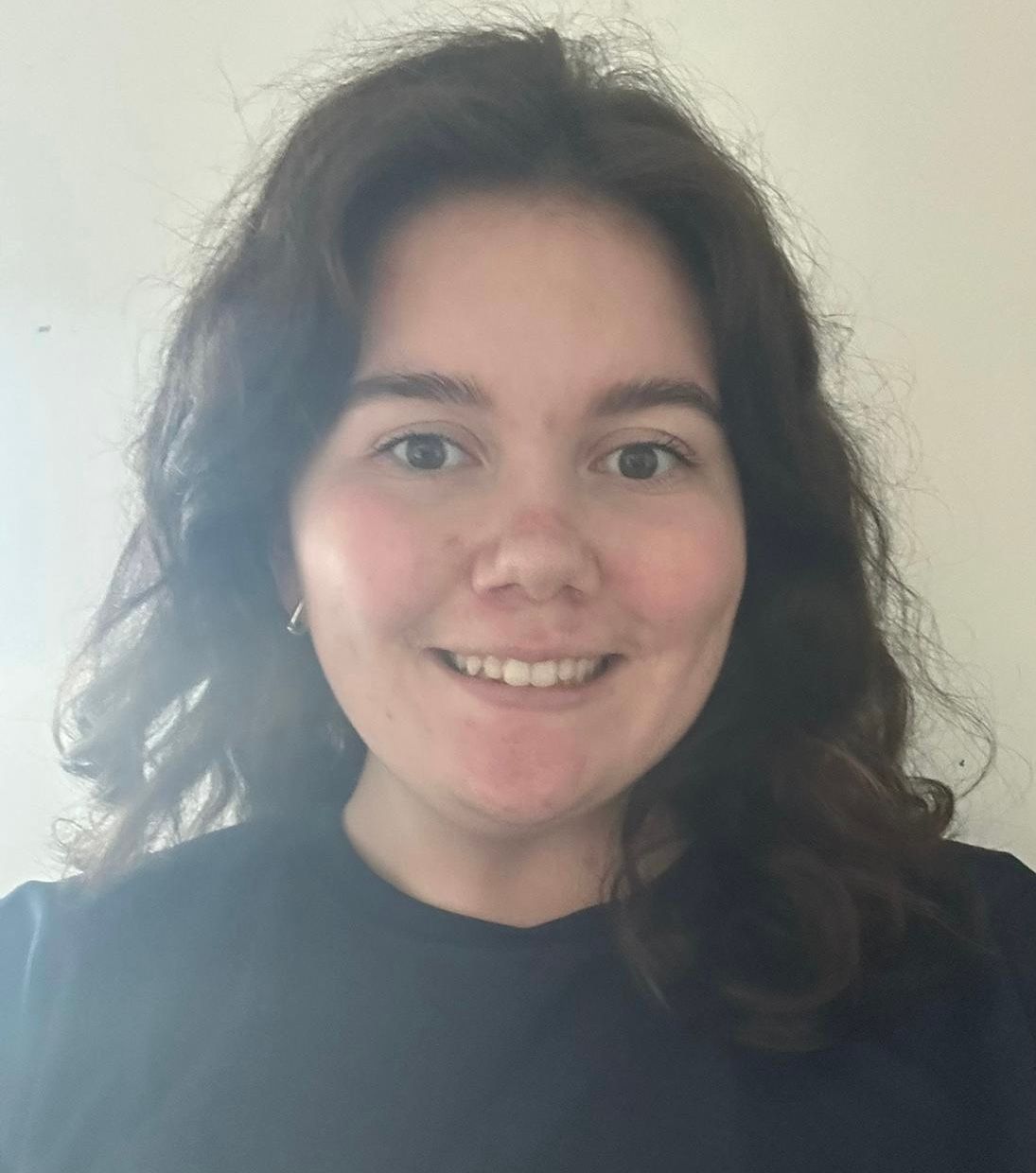 Smiling person with curly dark hair in a black shirt against a plain light wall