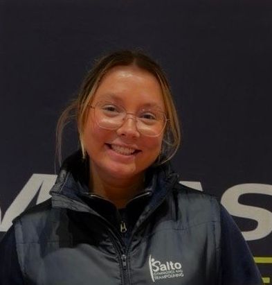 Smiling person in glasses and a black jacket standing before a dark wall with a Salto logo
