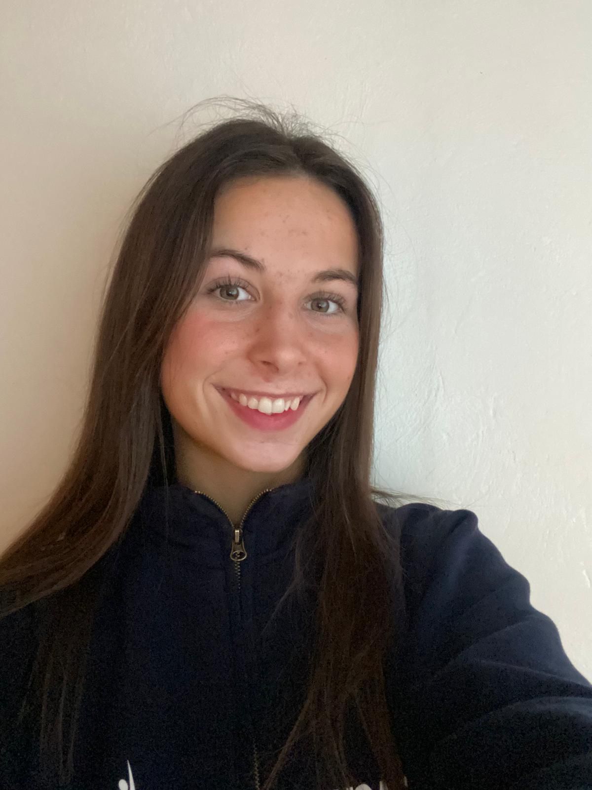 Smiling woman with long dark hair in a navy zip-up jacket against a white wall