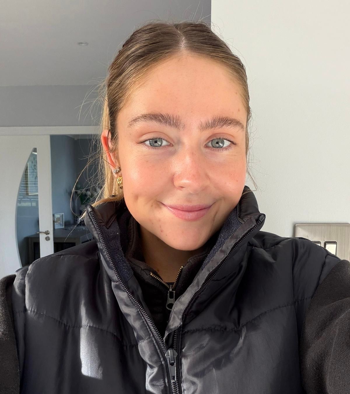 Smiling person in a black puffer jacket indoors, standing in front of a bright wall.