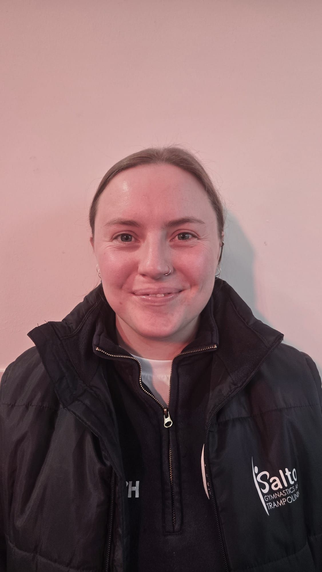 Person in a black jacket smiling against a plain pink wall.