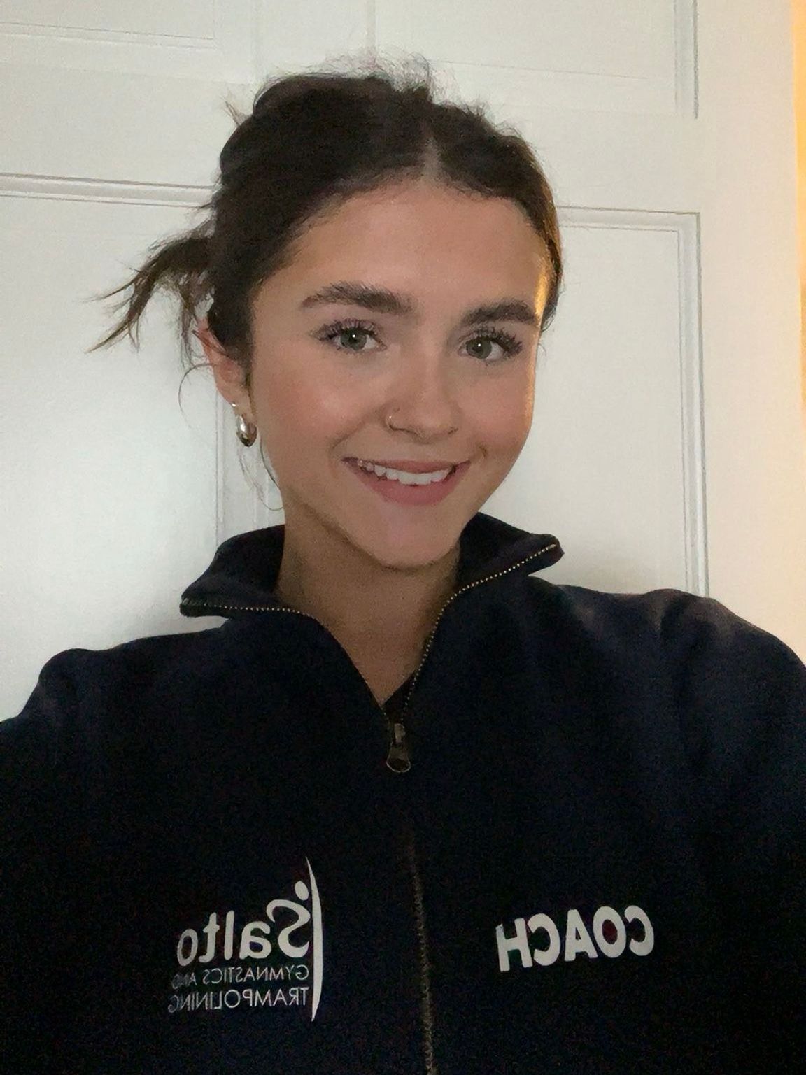 Smiling person in a black zip-up jacket with a white logo, standing indoors against a light door.