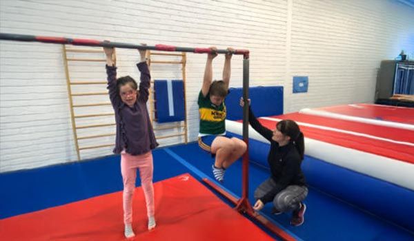 Children doing gymnastics on bars in a gym, with a coach assisting on a padded mat.