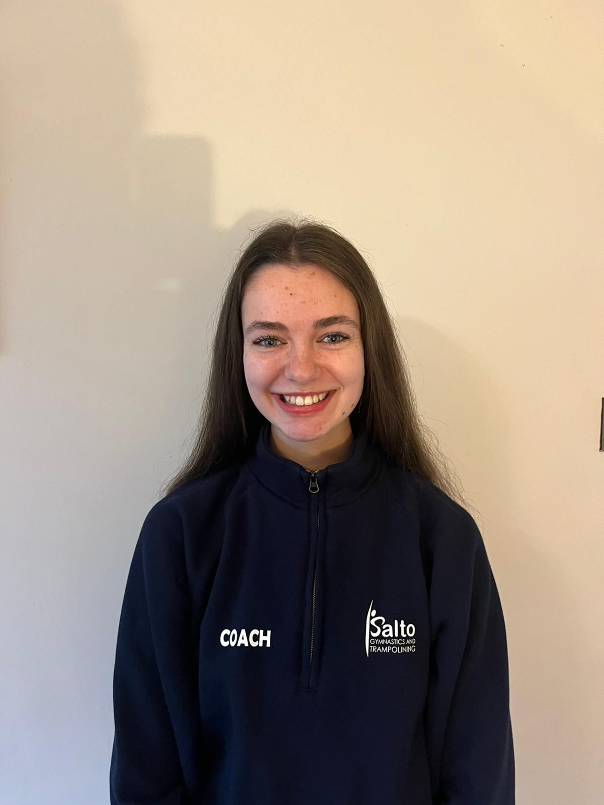 Smiling person in a navy zip-up jacket standing against a plain wall.