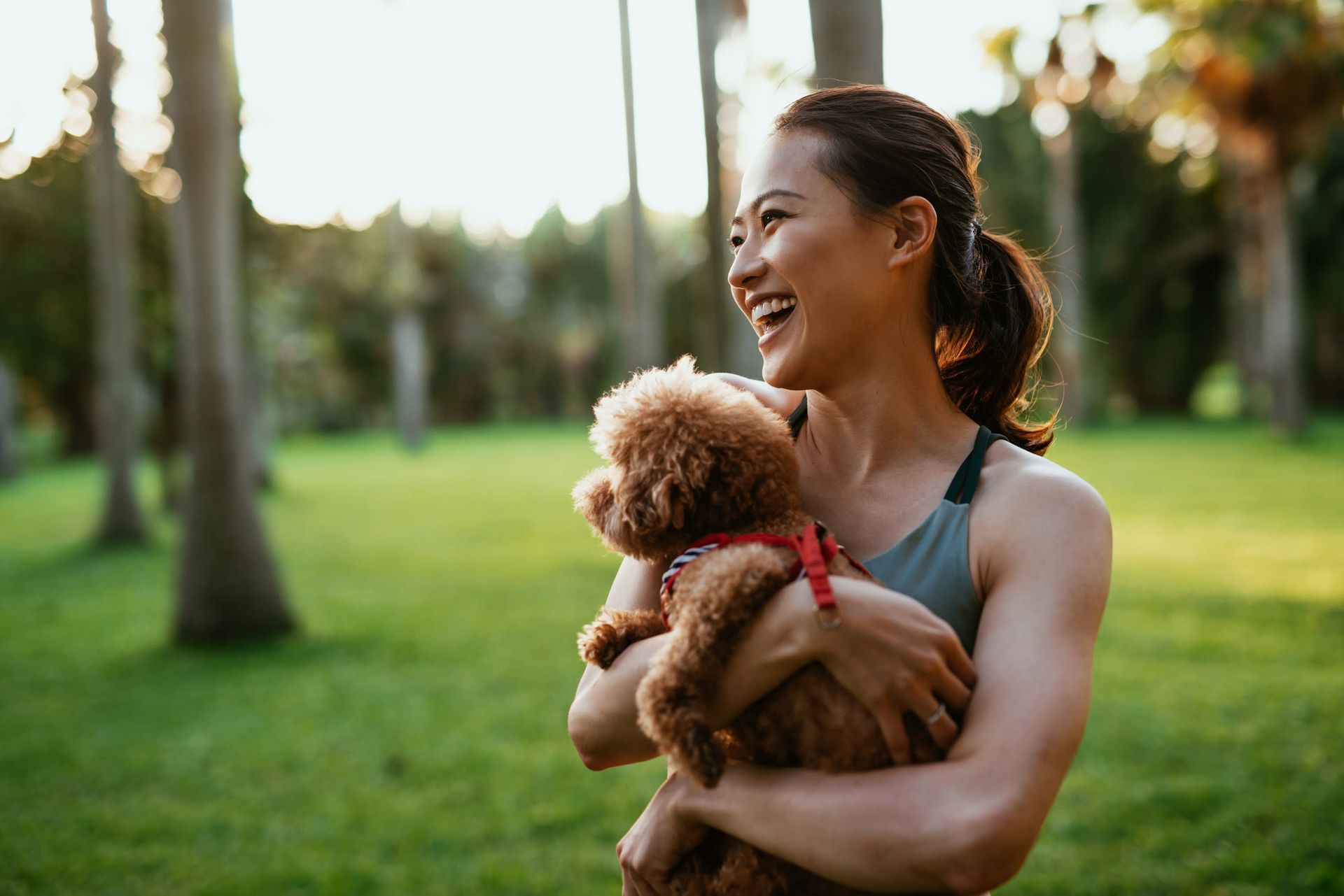 Smiling woman holding a small curly-haired dog in a sunny park