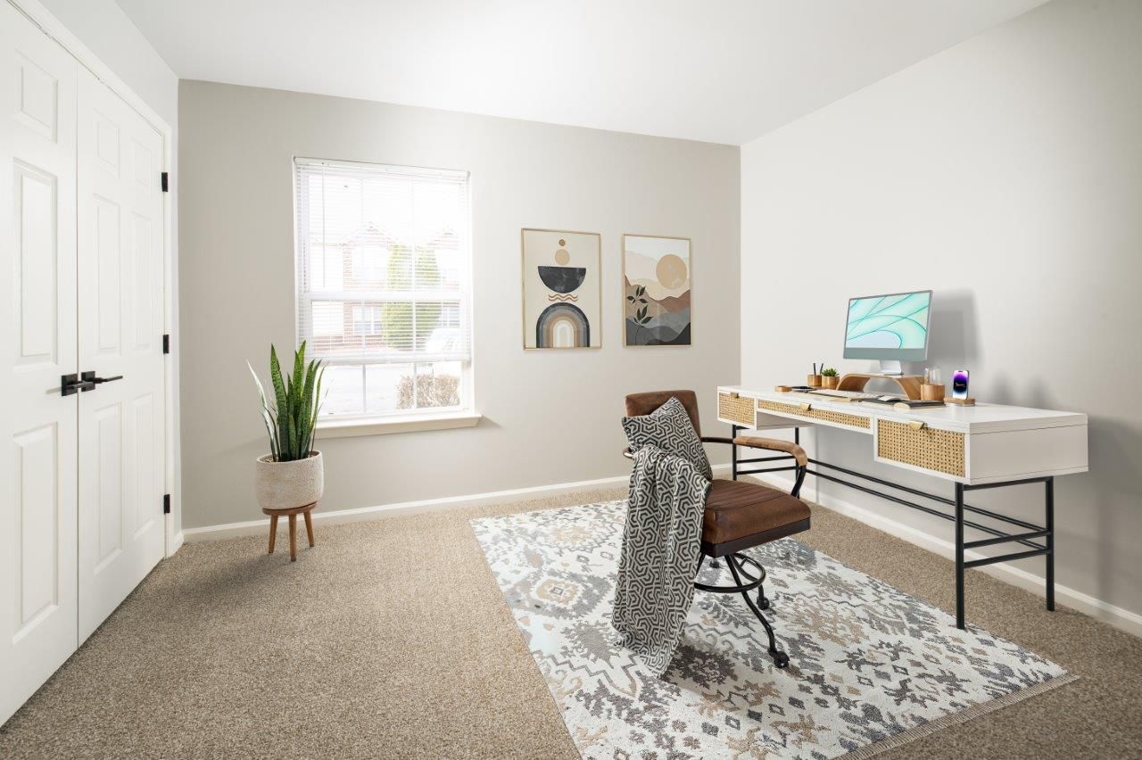 Bright home office with desk, chair, rug, window, and potted plant in a light beige room
