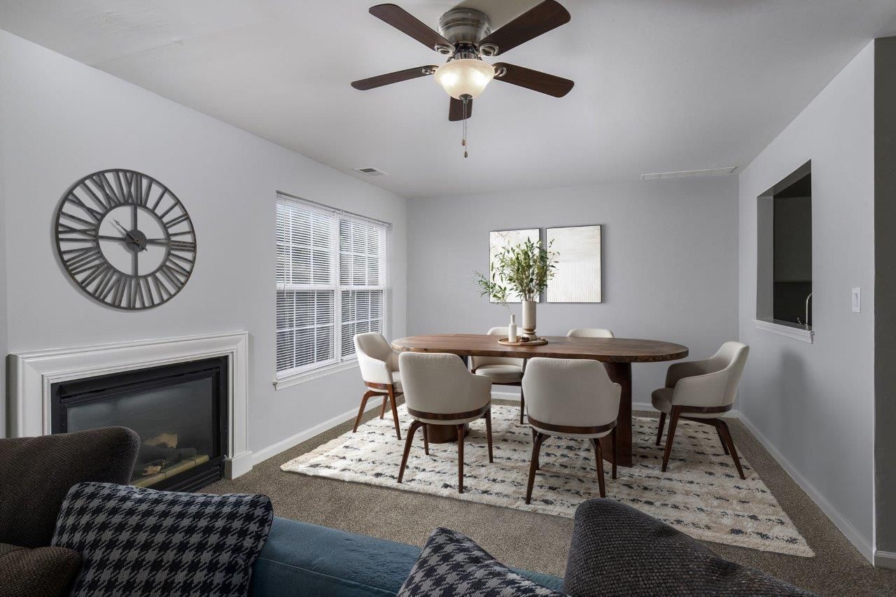 Bright dining room with round table, six chairs, fireplace, large wall clock, and ceiling fan
