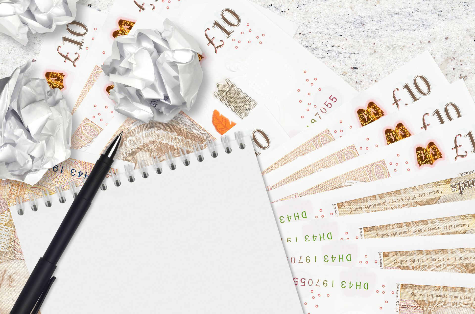 Notebook, pen, and crumpled papers on a background of ten-pound notes, suggesting financial planning or frustration.