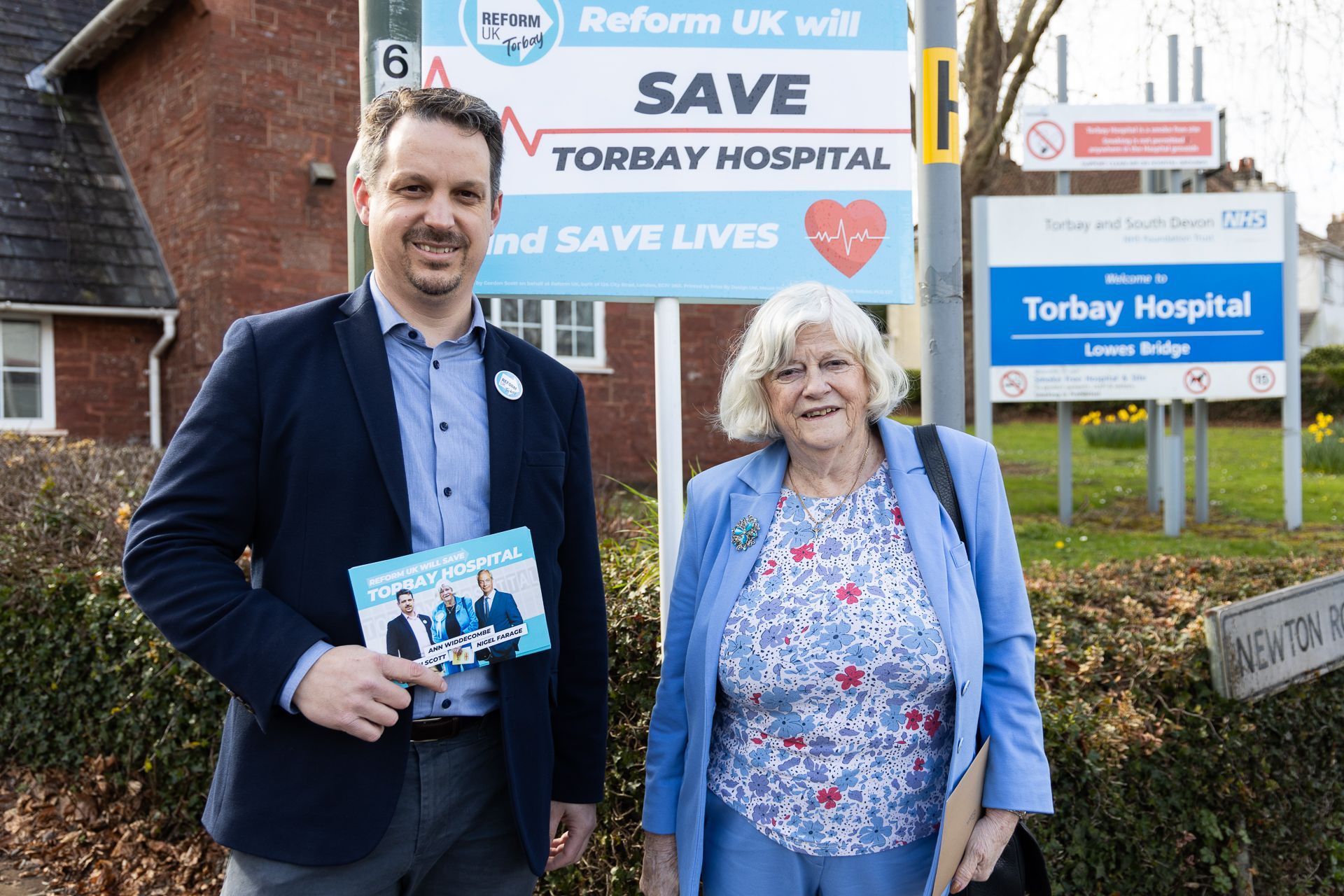 A man and a woman are standing in front of a sign that says save torbay hospital and save lives.