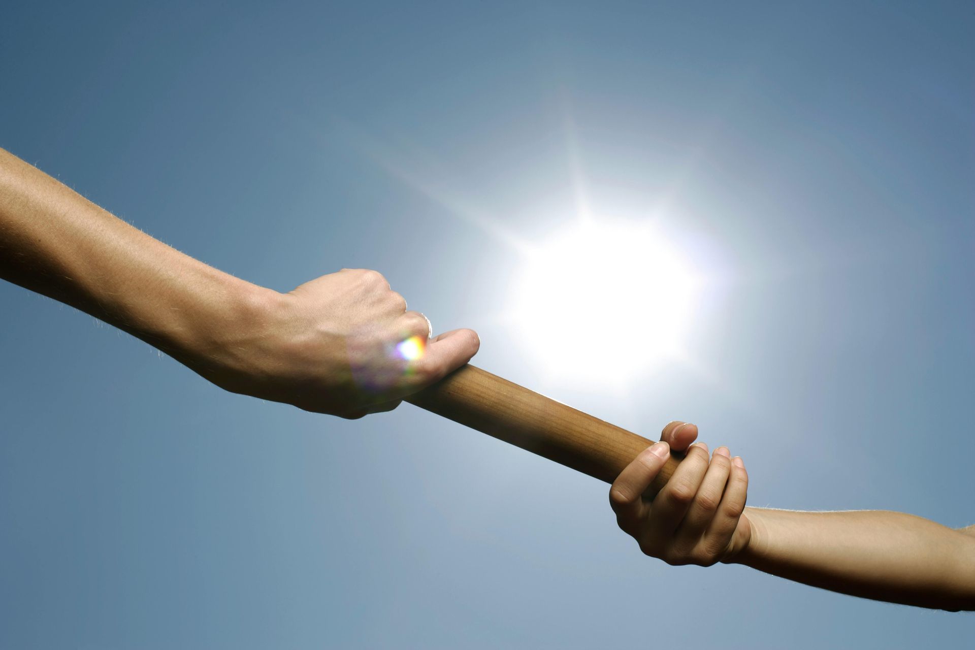 Two hands passing a baton against a bright sun and blue sky, symbolizing teamwork or relay race.