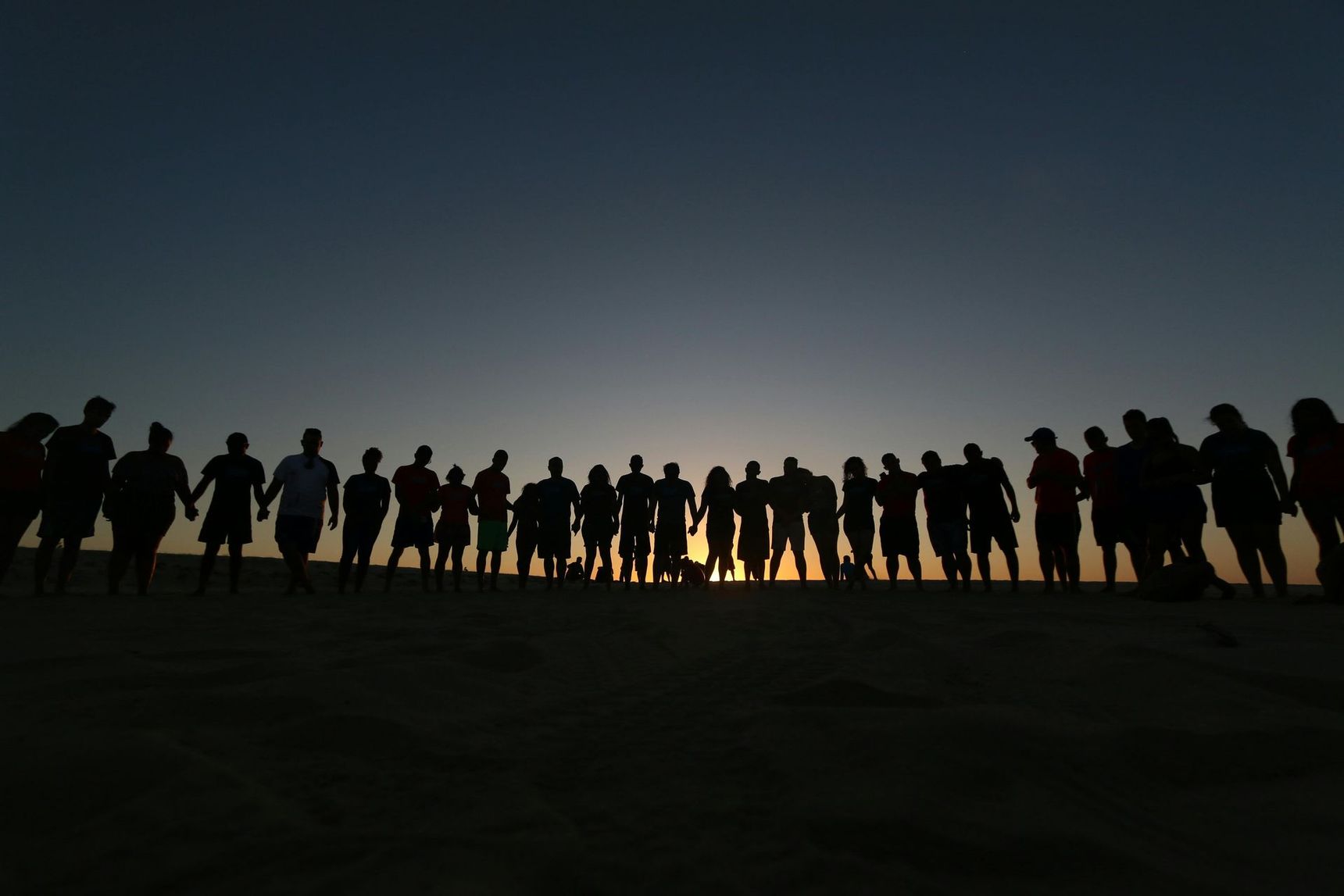 Un folto gruppo di persone in fila, che si tengono per mano, stagliano le proprie forme contro un tramonto luminoso su una duna di sabbia.