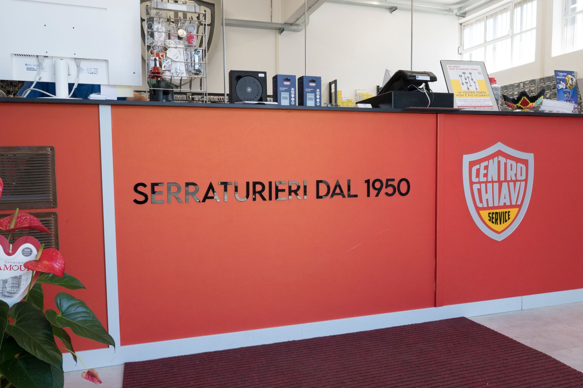 Bancone reception rosso con la scritta “SERRATURIFRI DAL 1950” e logo a scudo in un luminoso showroom adibito a officina.