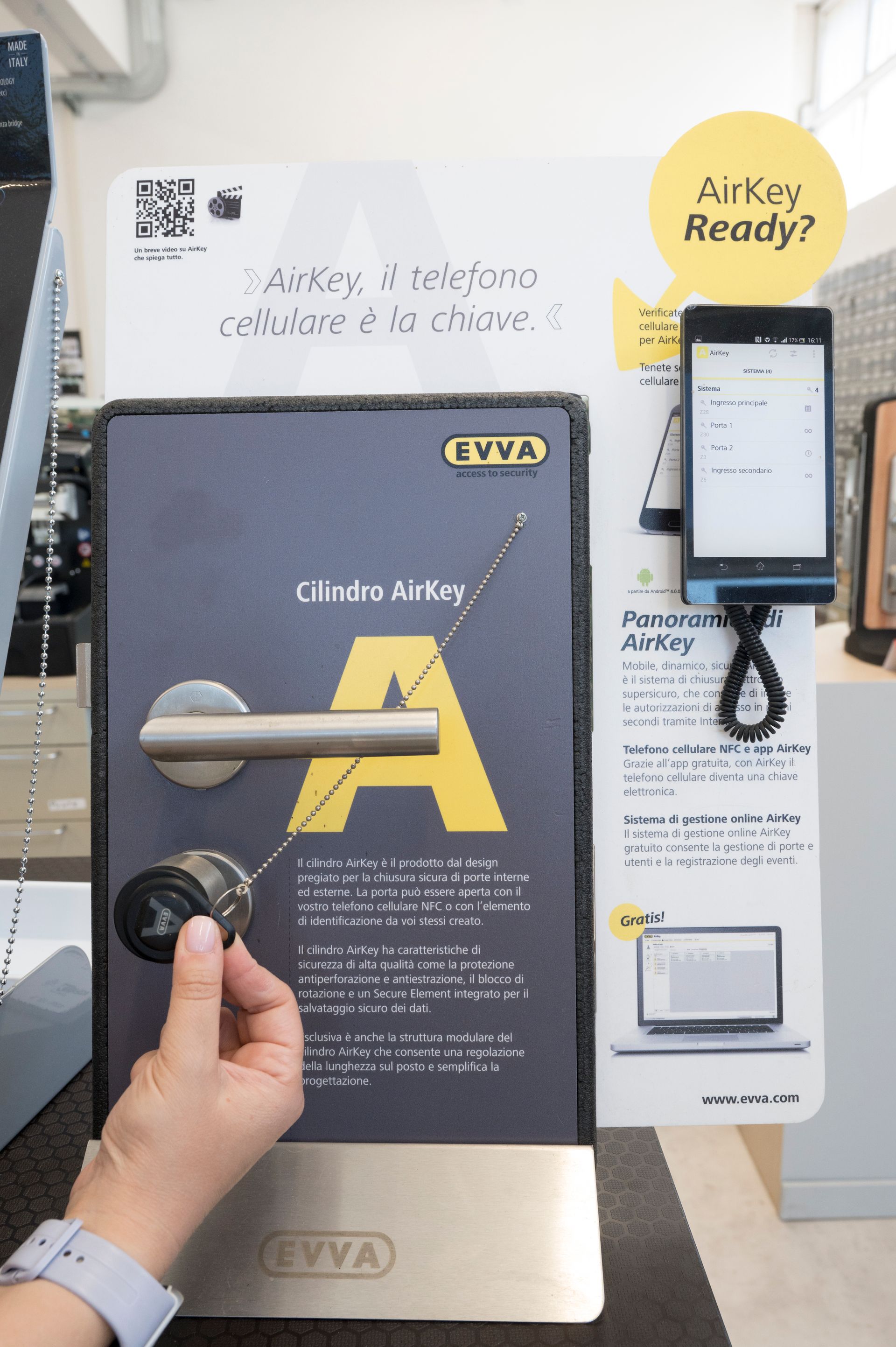 Una mano che regge un poster promozionale nero e oro del Club Airkey accanto a un chiosco touchscreen in un negozio.