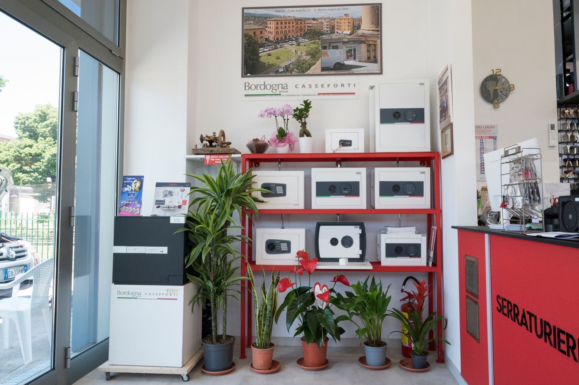 Interno luminoso del negozio con scaffali rossi, piante in vaso e piccoli elettrodomestici vicino a un ingresso in vetro.