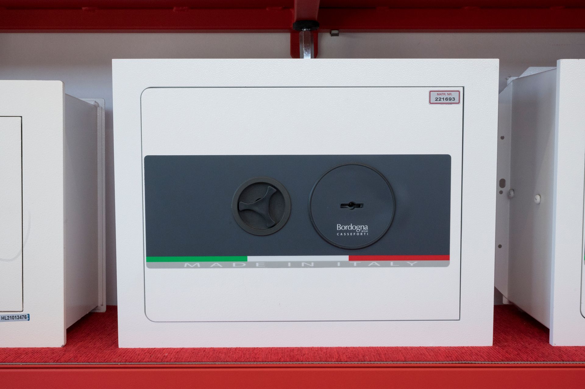 Cassaforte bianca con sportello grigio scuro su uno scaffale rosso, affiancata da altre casseforti simili.