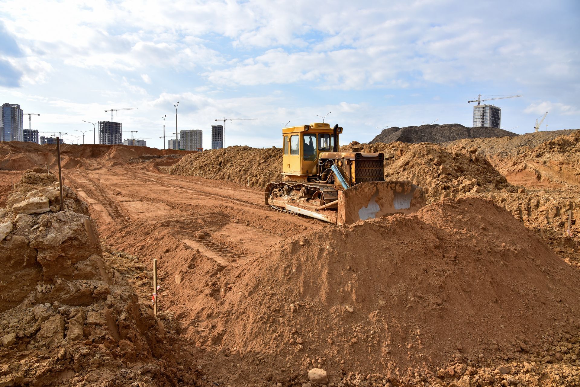 Bulldozer on a construction site, pushing dirt. Buildings and sky in background.
