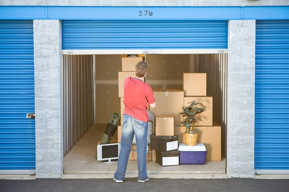 A Man In A Red Shirt Is Standing In A Storage Unit Filled With Boxes — Brisbane Moving & Storage In Acacia Ridge, QLD