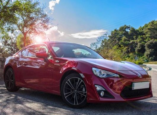 Red sports coupe parked outdoors on a sunny road with trees and blue sky in the background