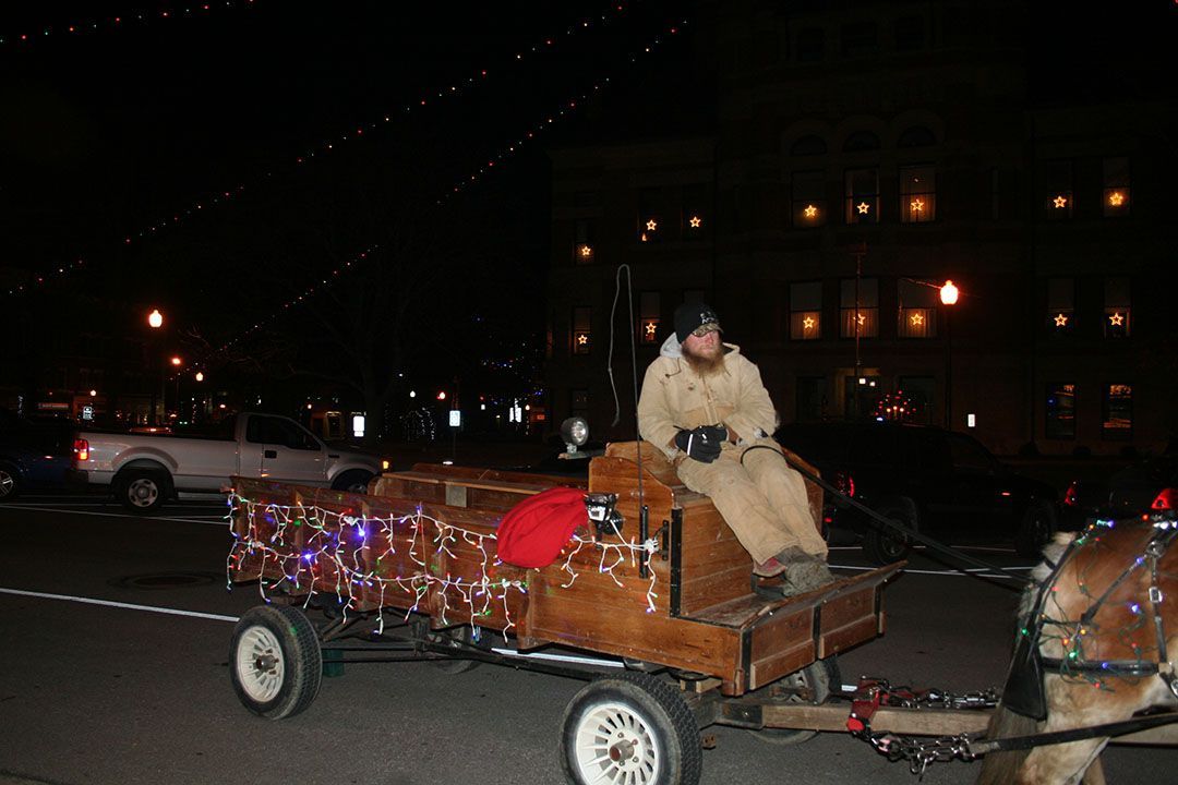 A person sits on a wooden, light-decorated wagon pulled by a horse at night in front of a lit building.