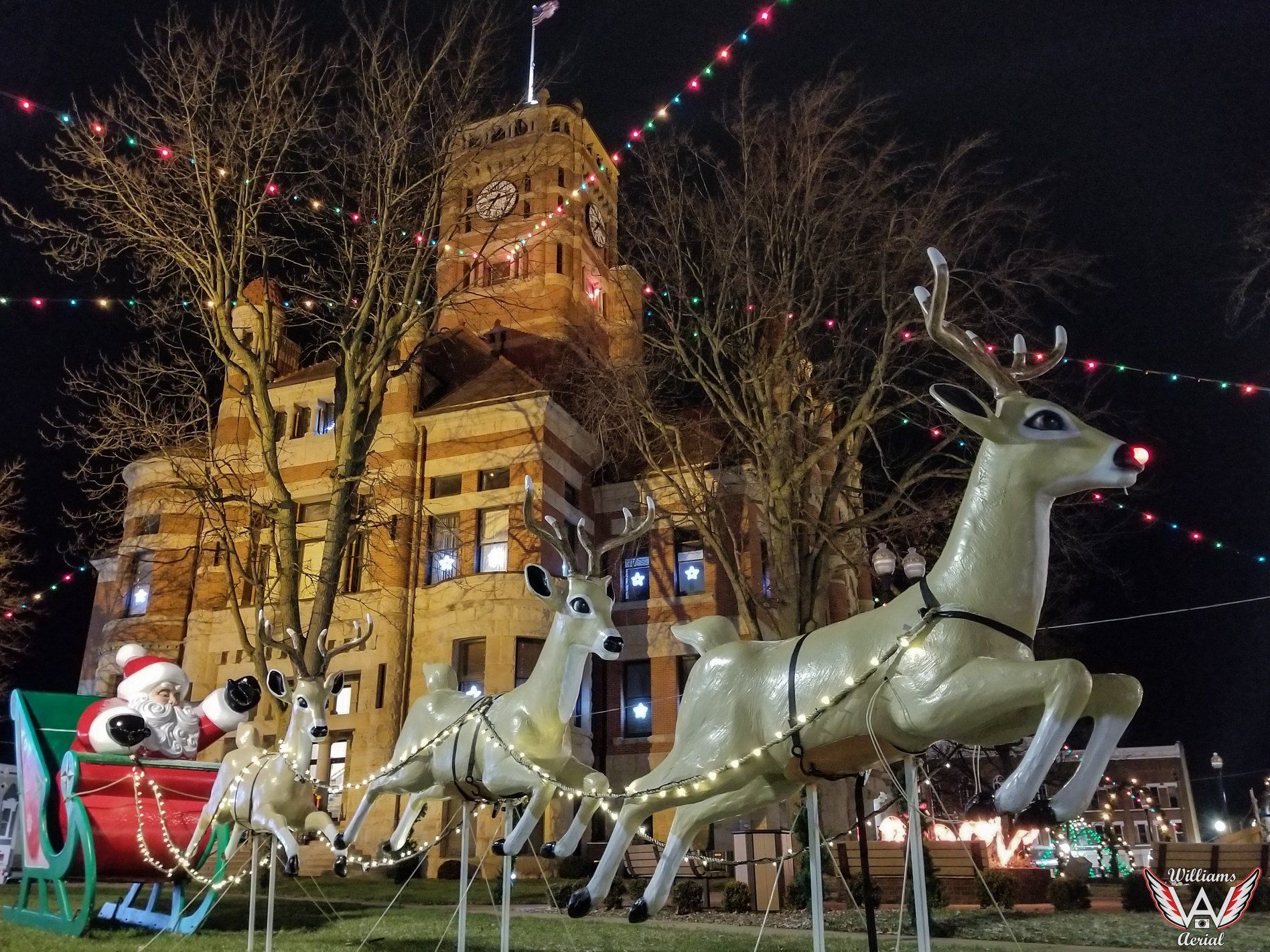 Santa sits in a red sleigh pulled by illuminated reindeer figures in front of a historic, lit clock tower at night.