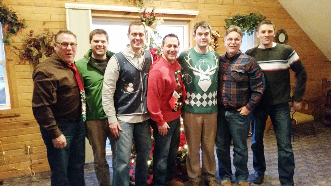 Seven people stand together in a rustic, wood-paneled room for a holiday-themed photo, some wearing festive sweaters.