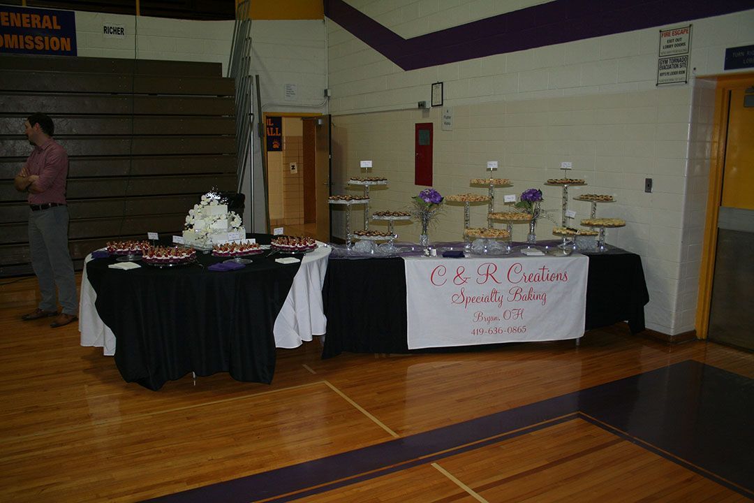 Tables with white and black cloths set up for an event in a gym, with a cake on one and a banner on the other.