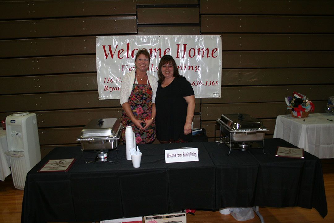 Two women stand smiling behind a black-covered buffet table with a "Welcome Home" banner hanging behind them in a gym.