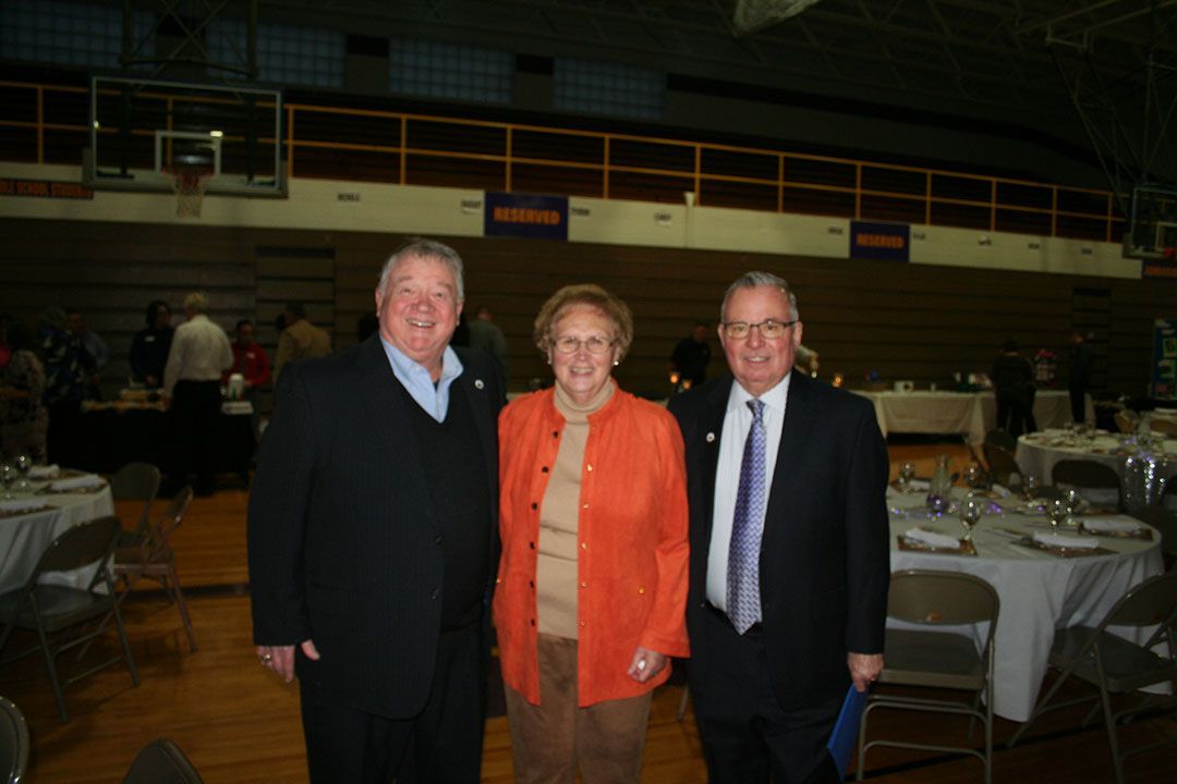 Three people stand in a gymnasium at a banquet, smiling for a photo with tables and chairs in the background.