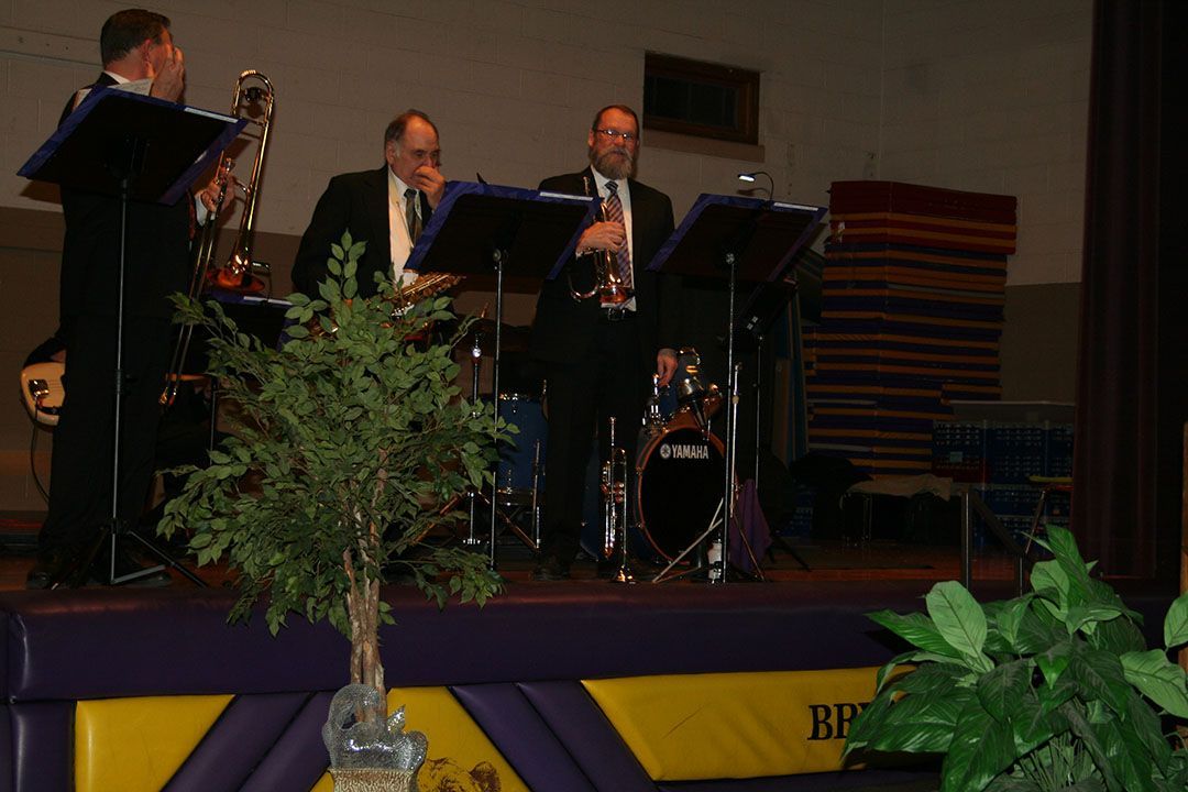 Three musicians in suits perform on a stage with music stands, a drum kit, and decorative plants in the foreground.